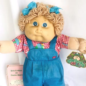 May include: A vintage Cabbage Patch Kids doll with blonde yarn hair and blue eyes. The doll is dressed in a red and white checked shirt and blue denim overalls. It has two blue bows and a hang tag.