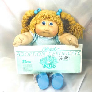 May include: A Cabbage Patch Kids doll with blonde yarn hair in pigtails, blue eyes, and dimpled cheeks. The doll wears a light blue gingham dress and holds an Official Adoption Certificate with the text Cabbage Patch Kids and Maggie Mae.