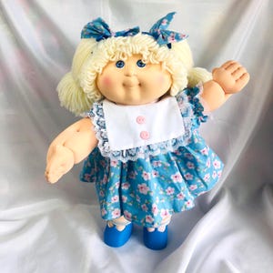 May include: A doll with blonde yarn hair styled in pigtails with blue floral bows, blue eyes, and rosy cheeks. The doll wears a blue dress with a pink floral pattern, a white bib collar with lace trim and two pink buttons, and blue shoes.