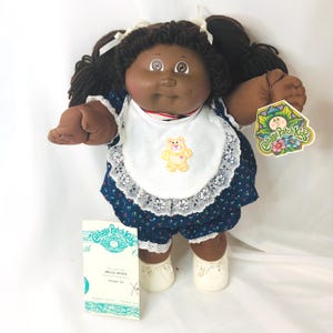 Puede incluir: Una muñeca Cabbage Patch Kids con cabello de lana marrón oscuro en coletas, piel morena y ojos marrones. La muñeca viste un vestido azul oscuro con estampado floral, un babero blanco con encaje y un oso amarillo bordado, y zapatos blancos.