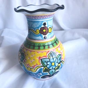Puede incluir: Un jarrón de cerámica pintado a mano con un borde con volantes. El jarrón presenta un diseño vibrante con patrones florales azules, amarillos y rojos. La base es redonda y el cuello es cilíndrico, con un borde blanco y negro. Es un objeto decorativo.