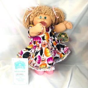 May include: A Cabbage Patch Kids doll with blonde yarn hair in pigtails, wearing a white dress featuring a colorful pattern of pumpkins and black cats. The doll has a pink pacifier in its mouth and comes with a Cabbage Patch Kids birth certificate.