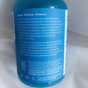K&ouml;nnte beinhalten: Eine blaue Flasche mit wei&szlig;em Text, der f&uuml;r Goli Ashwa Gummies wirbt. Das Etikett enth&auml;lt Informationen &uuml;ber die Vorteile des Produkts, wie Stressabbau und verbesserter Schlaf. Die Flasche hat auch ein Zertifizierungslogo.