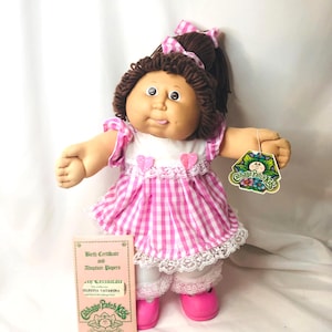 Puede incluir: Una muñeca Cabbage Patch Kids con cabello de lana marrón en coleta, vestida con un vestido de cuadros vichy rosa y blanco con ribete de encaje, dos pequeños corazones rosas y pololos blancos. La muñeca lleva zapatos rosas y una etiqueta.