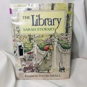 May include: A hardcover book titled "The Library" by Sarah Stewart, with illustrations by David Small. The cover features a whimsical illustration of a person pulling a cart of books, a black cat, and a white picket fence.