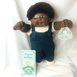 Puede incluir: Una muñeca Cabbage Patch Kids de piel oscura, ojos marrones y pelo corto rizado de color marrón oscuro. La muñeca lleva una camisa color crema bajo un peto vaquero azul oscuro y zapatos blancos. Se incluyen una etiqueta y un certificado.