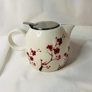 Może przedstawiać: Biały ceramiczny czajnik z pokrywką i uchwytem ze stali nierdzewnej. Czajnik jest ozdobiony czerwonym wzorem gałęzi kwiatowych. Wylewka znajduje się po prawej stronie czajnika.