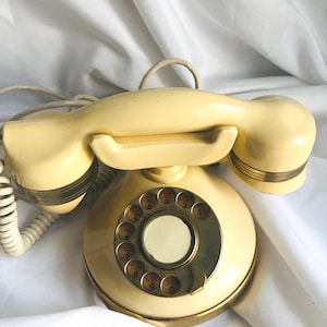 May include: Cream-colored vintage rotary phone with a circular dial and brass accents. The handset rests on the base, connected by a coiled cord. The dial features numbers and letters. A classic piece of communication technology.