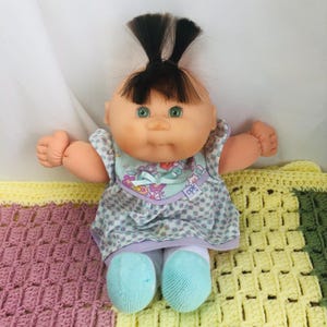 Puede incluir: Una muñeca Cabbage Patch Kids vintage con ojos verdes, cabello negro y marrón y un vestido con estampado floral. La muñeca tiene un babero azul claro con un diseño floral y está sentada sobre una colorida manta de ganchillo. Los pies de la muñeca son de color azul claro.