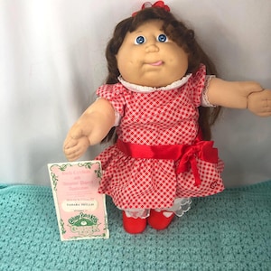 Op de afbeelding: Een Cabbage Patch Kids pop met bruin haar, blauwe ogen en een rood-wit geruite jurk. De pop heeft een rode strik in haar haar en om haar taille. Ze houdt een certificaat vast.