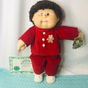 Puede incluir: Muñeca Cabbage Patch Kids, Aristotle Terry, vestida con un atuendo de pana rojo con un motivo de oso de peluche. La muñeca tiene cabello castaño, mejillas rosadas y zapatos blancos. Se incluye un certificado de nacimiento.