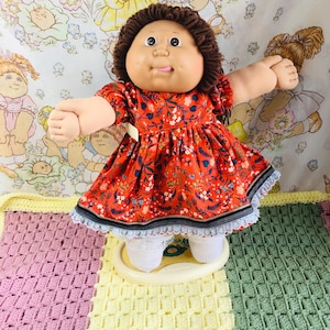 Può includere: Bambola Cabbage Patch Kid vintage con capelli castani e un vestito rosso floreale. La bambola ha un viso rotondo, guance rosee e si trova su una base color crema. L'abito ha un bordo in pizzo bianco.