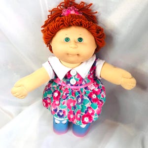 Könnte beinhalten: Eine Puppe mit wollartigem, kastanienbraunem Haar, das mit einer rosa Schleife verziert ist, grünen Augen und einem runden Gesicht. Die Puppe trägt ein weißes Kleid mit Kragen und einem floralen Muster in Pink, Lila und Grün sowie zwei Knöpfen.