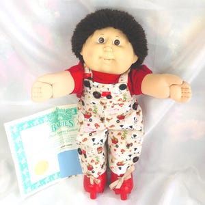 May include: A Cabbage Patch Kids doll with dark brown yarn hair, brown eyes, and a dimpled face. The doll wears a red short-sleeved shirt under beige overalls featuring a farm-themed print with tractors and animals, paired with red shoes.