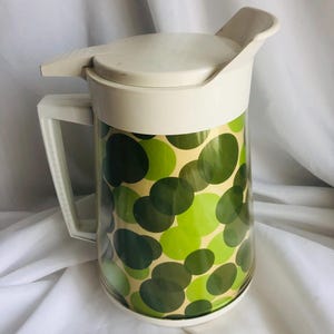 Op de afbeelding: Een vintage kan met een witte deksel en handvat. De body van de kan is versierd met een patroon van groene en olijfkleurige cirkels. De plastic kan heeft een capaciteit van ongeveer 1,9 liter.
