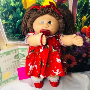 Può includere: Una bambola Cabbage Patch Kids con capelli di lana marrone a codini, che indossa un vestito rosso con un motivo bianco di figure e stelle, bordo in pizzo bianco e scarpe rosse. La bambola ha un occhio blu e uno scuro e un ciuccio rosso in bocca.