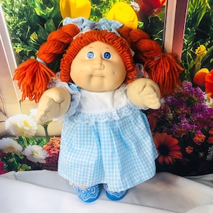 Op de afbeelding: Een pop met fel oranje wollen haar in twee dikke vlechten, blauwe ogen en kuiltjes in de wangen. De pop draagt een lichtblauw met wit geruite jurk met een wit lijfje, kanten afwerking bij de kraag en bijpassende blauwe schoenen met kant.