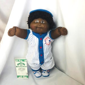 May include: A Cabbage Patch Kids doll with dark skin and black yarn hair, wearing a blue and white pinstriped baseball uniform with red buttons and a blue cap. The doll has a birth certificate labeled Albert Quincy and white sneakers.