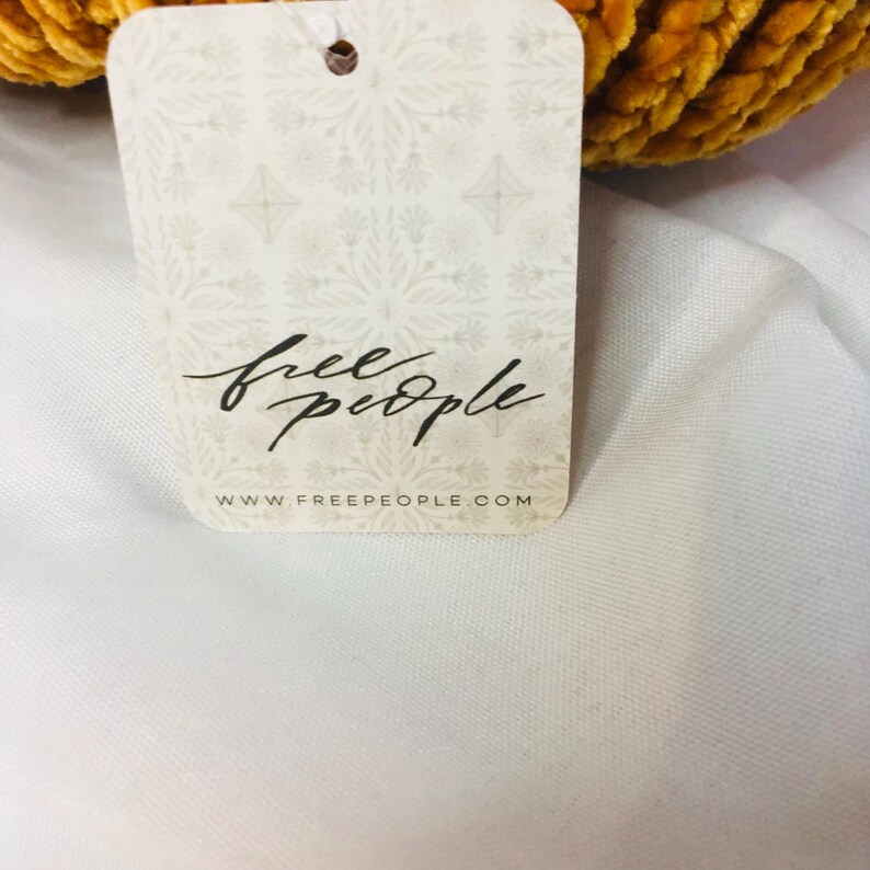 Peut inclure: Une &eacute;tiquette cr&egrave;me avec les mots "free people" en &eacute;criture cursive. L'&eacute;tiquette pr&eacute;sente un motif floral et un petit trou en haut. L'adresse du site Web www.freepeople.com est imprim&eacute;e en bas.