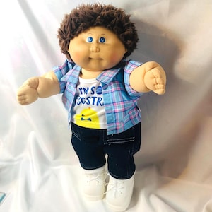 Könnte beinhalten: Eine Stoffpuppe mit braunen Wollhaaren und blauen Augen, bekleidet mit einem weißen T-Shirt mit der Aufschrift I'M SO EGGSTRA und einem gelben Küken-Motiv, einem blau-rosa karierten Hemd, einer dunkelblauen Jeanshose und weißen Schnürschuhen.