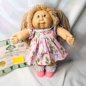 May include: A Cabbage Patch Kids doll with tan yarn hair, green eyes, and dimples, wearing a pink dress with a strawberry print, white lace-trimmed socks, and pink shoes. Beside the doll is an official adoption certificate with printed text.