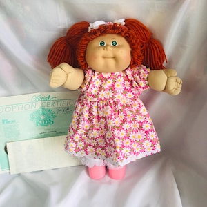 May include: A Cabbage Patch Kids doll with auburn yarn hair in pigtails, green eyes, and a light complexion. The doll wears a pink dress with a white daisy pattern, white lace-trimmed bloomers, and pink shoes, accompanied by an adoption certificate.
