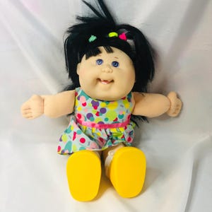 May include: A doll with black hair styled in a ponytail with small green, yellow, and pink hair ties. The doll has blue eyes, a smiling mouth with one visible tooth, and wears a light blue dress featuring a colorful polka dot pattern.