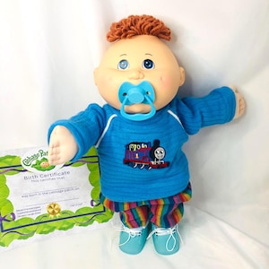Pode incluir: Uma boneca Cabbage Patch Kids com cabelo de lã laranja, olhos azuis e uma chupeta azul clara. A boneca veste uma camisa azul de manga comprida com um gráfico do Thomas e seus Amigos, calças listradas multicoloridas e sapatos azul claros, acompanhada de uma certidão de nascimento.
