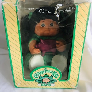 May include: A Cabbage Patch Kids bank doll in its original packaging. The doll has black hair, green accents, and is holding a pink pig. The packaging is yellow and green with the Cabbage Patch Kids logo and the word "BANK".