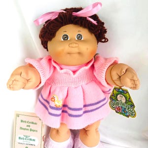 May include: A Cabbage Patch Kids doll with brown yarn hair tied with a pink ribbon, brown eyes, and rosy cheeks. The doll wears a pink knit dress with a yellow duck graphic and purple stripes, and includes a tag and a birth certificate document.