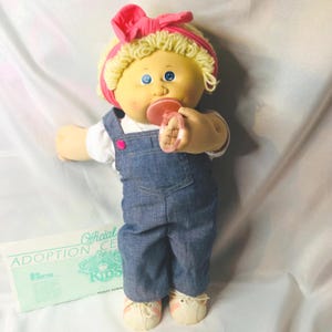 Op de afbeelding: Een Cabbage Patch Kids pop met blond wollen haar, een roze hoofdband en blauwe ogen. De pop draagt een wit shirt, een blauwe spijkerbroek met een roze knoop en witte sneakers met roze strepen. Een roze fopspeen zit in de mond. Een officieel adoptiedocument is inbegrepen.