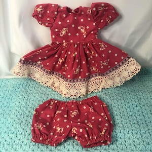 May include: A doll's dress and matching bloomers set. The red fabric features white polka dots and floral patterns. The dress has short, puffed sleeves and a lace trim along the hem. The bloomers have an elastic waistband and cuffs.