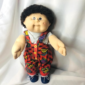 May include: A doll with dark brown yarn hair and a round face featuring a small dimple on the left cheek. The doll wears a light gray short-sleeved shirt under red and black plaid overalls with yellow buttons and dark blue shoes with white laces.