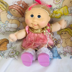 Puede incluir: Una muñeca vintage de Cabbage Patch Kids con cabello rubio, ojos azules y pecas. La muñeca lleva un vestido rosa con adornos dorados y calcetines de encaje blanco. Se ven los zapatos rosas de la muñeca. La muñeca está sentada sobre una tela estampada floral.