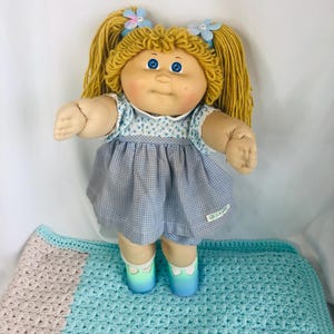 Può includere: Una bambola con capelli di lana in due codini biondi, occhi blu e guance rosate. La bambola indossa un corpetto con stampa floreale blu e bianca, una gonna a quadretti blu e bianca e scarpe sfumate blu e bianche con calzini bianchi.