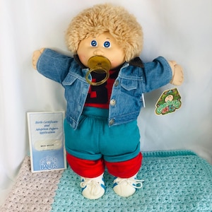 Könnte beinhalten: Eine Cabbage Patch Kids Puppe mit blondem lockigem Haar und blauen Augen, bekleidet mit einer blauen Jeansjacke über einem rot-schwarzen Oberteil, einer türkisfarbenen Hose mit roten Bündchen und weißen Turnschuhen. Ein gelber Schnuller ist im Mund.