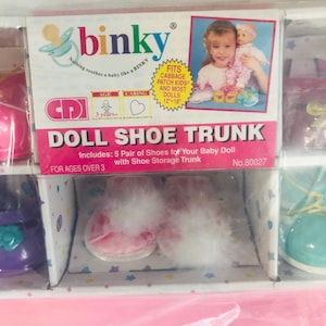 Puede incluir: Un baúl de zapatos de muñeca con la marca "binky" y el texto "Nothing soothes a baby like a BINKY". La caja incluye cinco pares de zapatos para muñecas y es para mayores de 3 años. La caja tiene una foto de una niña sosteniendo una muñeca.