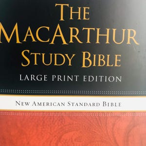 Op de afbeelding: De MacArthur Study Bible, Large Print Edition, met de tekst "New American Standard Bible" onderaan. De titel is in gouden letters op een zwarte achtergrond, met een rood en wit patroon aan de onderkant.