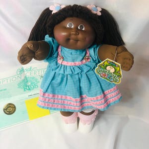 May include: A Cabbage Patch Kids doll with dark brown yarn hair styled in pigtails with pink and blue flower hair clips. The doll wears a teal plaid dress with pink trim and a ruffled bodice, white socks with pink ruffles, and white shoes.