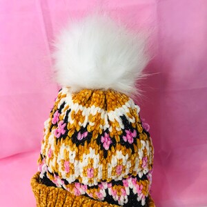 Peut inclure: Un bonnet d'hiver tricot&eacute; avec un pompon en fausse fourrure blanche. Le bonnet pr&eacute;sente un motif de couleurs or, blanc, noir et rose. Le chapeau est fait d'une mati&egrave;re douce et textur&eacute;e, parfaite pour le froid.