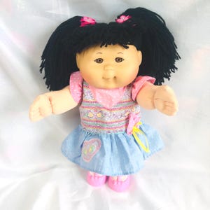 May include: A doll with black yarn hair styled in two pigtails with pink flower accents. The doll has brown eyes and wears a dress with a pink polka-dot bodice, a patterned midsection, and a light blue skirt featuring a heart-shaped patch.