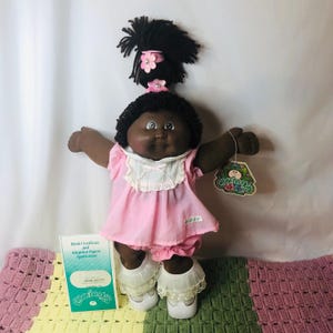 Könnte beinhalten: Eine Cabbage Patch Kids Puppe mit dunkler Haut und schwarzem Wollhaar, das zu einem hohen Pferdeschwanz mit zwei rosa Blumen-Haaraccessoires gebunden ist. Die Puppe trägt ein rosa Kleid mit weißem Spitzenkragen, rosa Pumphosen und weiße Schuhe.