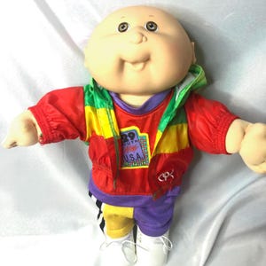 May include: A bald doll with a round face and brown eyes, wearing a multi-colored jacket with red, yellow, and green panels. The jacket features a patch with the text 29 SOUTH U.S.A. The doll wears purple and yellow pants and white shoes.