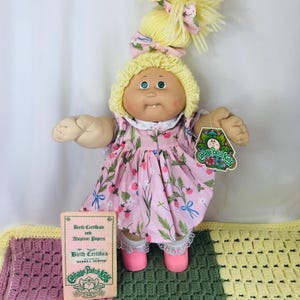 Puede incluir: Una muñeca Cabbage Patch Kids con cabello de lana amarilla, ojos verdes y un diente. La muñeca viste un vestido rosa con estampado de fresas y lazos azules, calcetines con borde de encaje blanco y zapatos rosas. Incluye certificado.