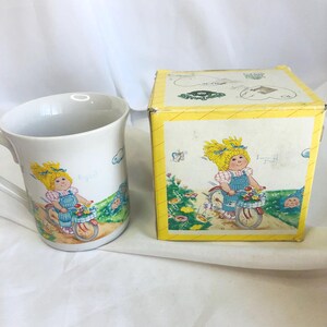 Puede incluir: Taza de cerámica blanca con asa, con una ilustración de dibujos animados de una niña montando en bicicleta con una cesta de flores. Una caja a juego con la misma ilustración está al lado de la taza.
