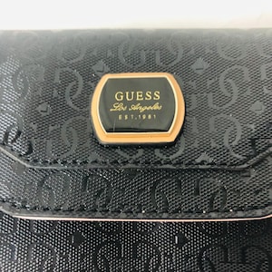 Puede incluir: Cartera negra con un patrón en relieve repetido. Una placa dorada con el texto "GUESS Los Angeles EST. 1981" está centrada en la parte delantera. La cartera tiene un ribete rosa.