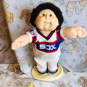 May include: A soft-sculpture doll with dark brown yarn hair and a round face, wearing a white, red, and blue baseball jersey with the text SOX printed on the chest and a Major League Baseball logo on the sleeve, paired with matching white pants.