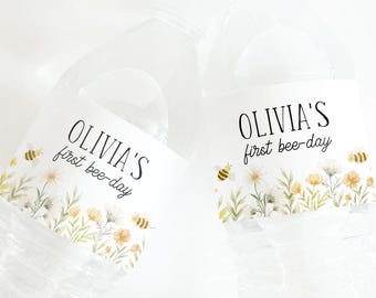 Op de afbeelding: Doorzichtige plastic waterflessen met etiketten met de tekst "OLIVIA'S first bee-day". De etiketten hebben een bloemenontwerp met gele bloemen, groene bladeren en kleine bijenillustraties. De flessen zijn gedeeltelijk gevuld met water.