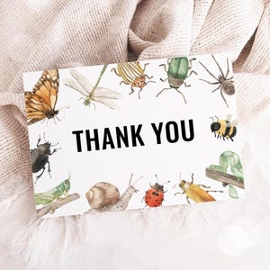 May include: A white card with the words "THANK YOU" in bold black letters. The card is decorated with watercolor illustrations of various insects, including a butterfly, dragonfly, bee, and ladybug. The card is set against a soft, neutral-colored background.
