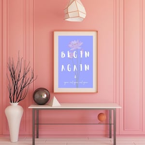 Può includere: Una stampa incorniciata con le parole "BEGIN AGAIN" in bianco su sfondo lilla chiaro, con un'illustrazione floreale. La stampa è esposta su un tavolo bianco contro un muro rosa. Sono visibili anche un vaso bianco con rami e una lampada geometrica.